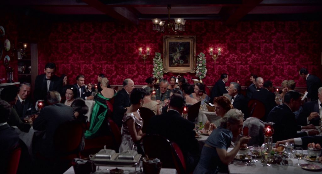 Wide shot of a dining room with red velvet walls. Everyone is dressed in greys, blacks, and blues except one woman in emerald green. Color palettes